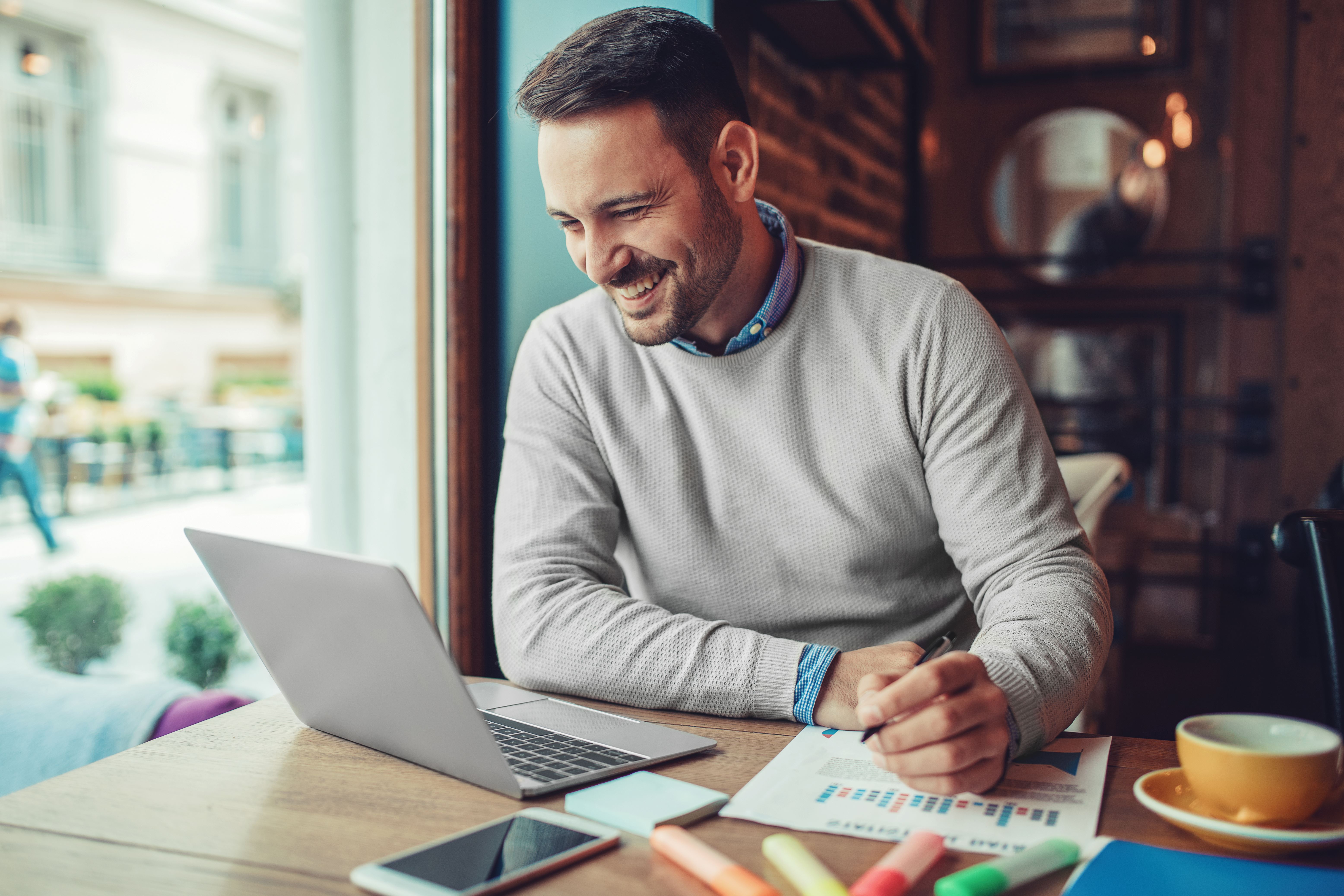 Man in grijze trui, lachend en aan het werk in café, kijkend naar laptop