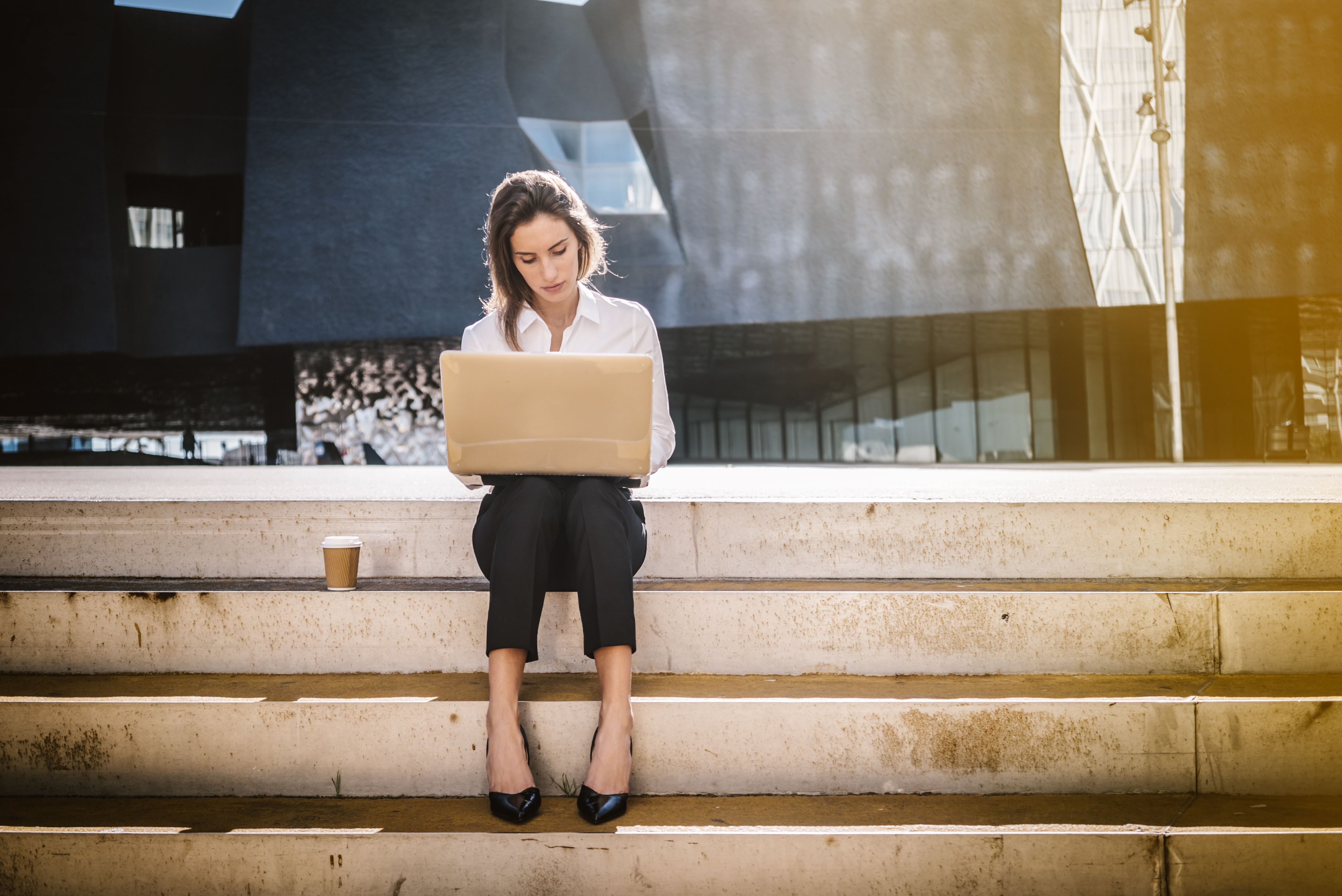 Een vrouw zit op een trap met haar laptop op schoot. Naast haar staat een kopje koffie.