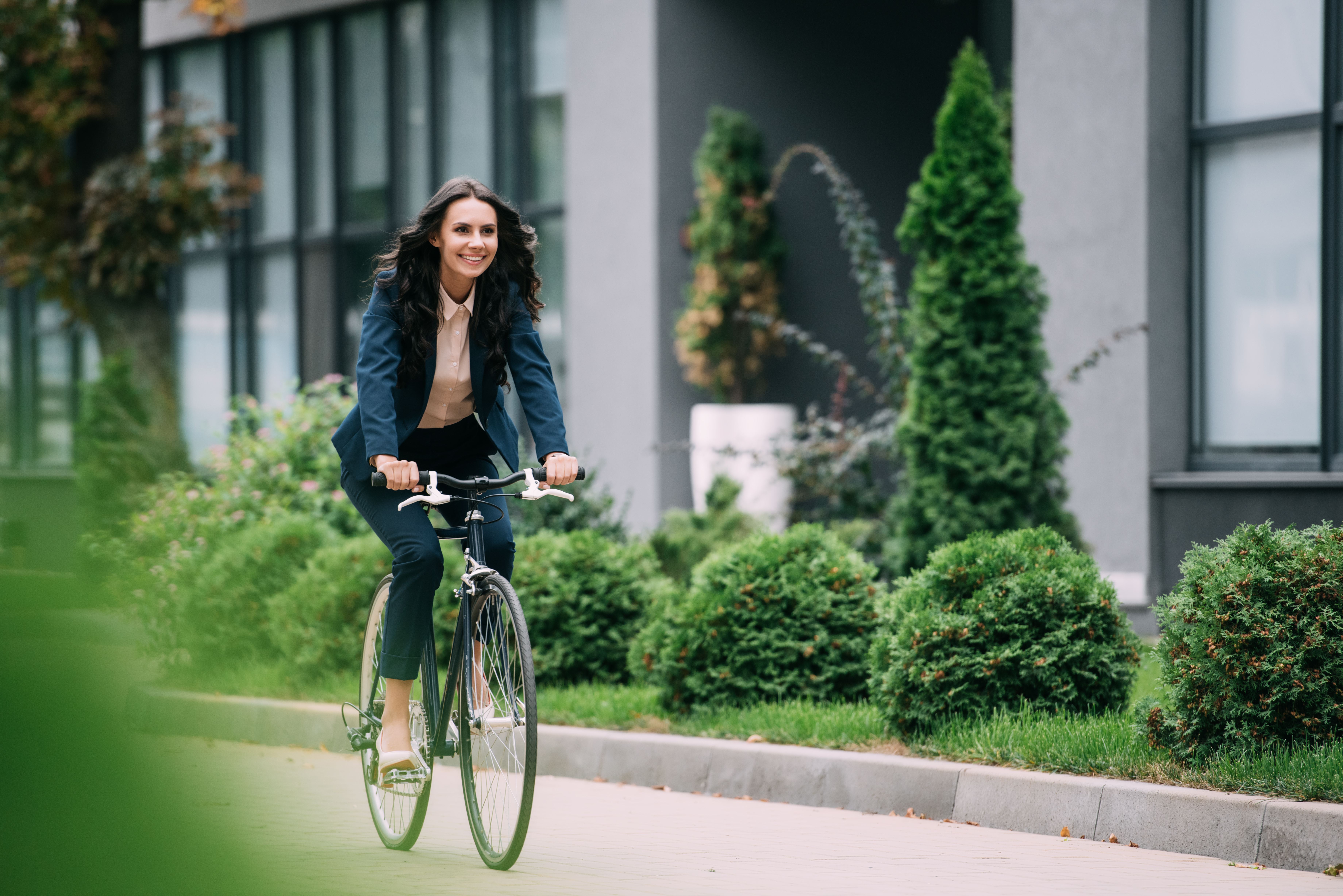 Een vrouw in zakelijke kleding fietst op het fietspad, naast het pad ligt een grasveldje met bosjes.