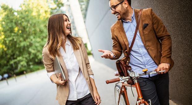 Een man en een vrouw lopen over straat, de man heeft een rode fiets in de hand. De twee mensen lachen naar elkaar