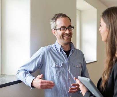 Een man met een blauwe blouse en een bril staat te praten met een vrouw met donker bruine haren.