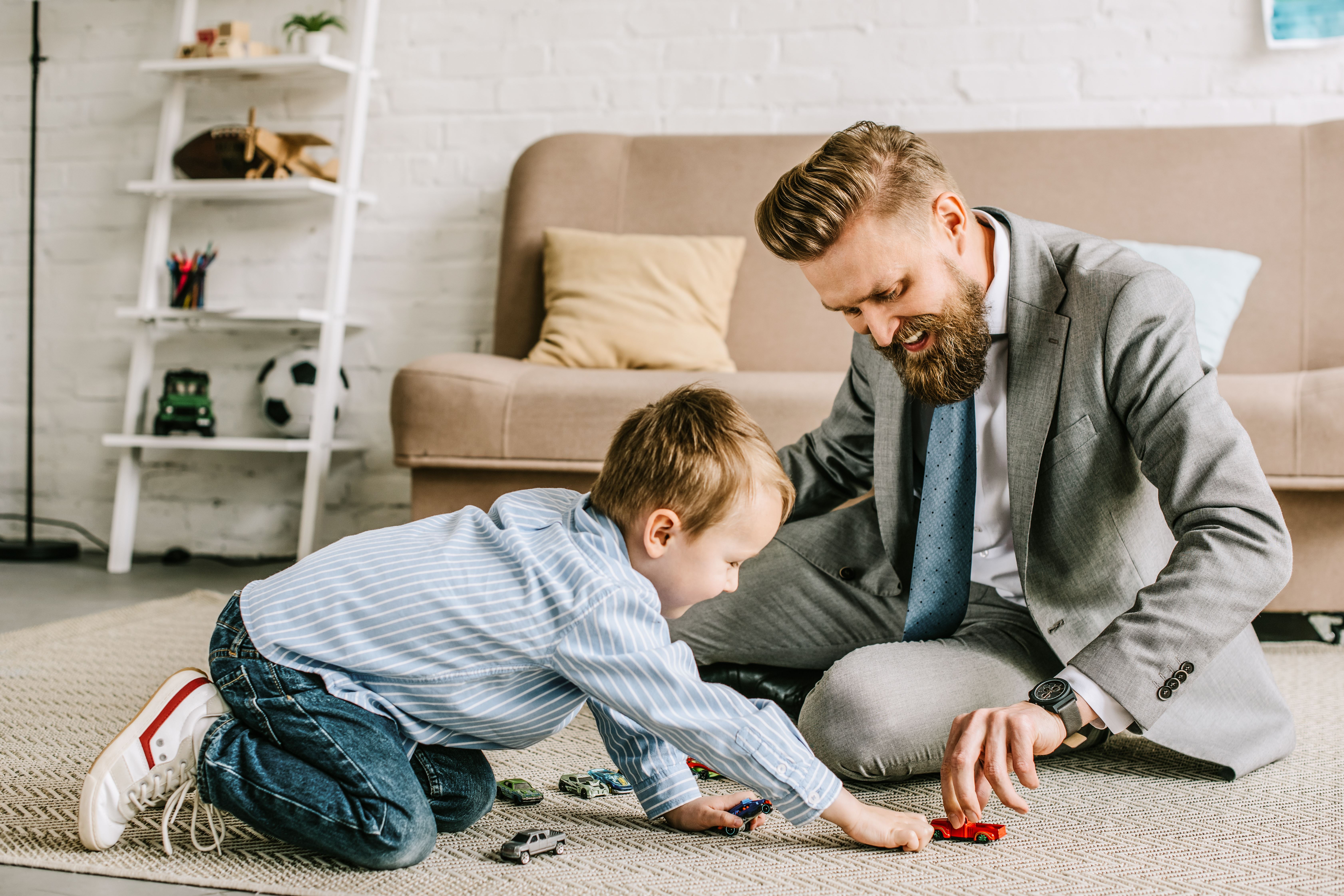 Een man in pak speelt met zijn zoontje op de grond in de woonkamer met een speelgoed auto.